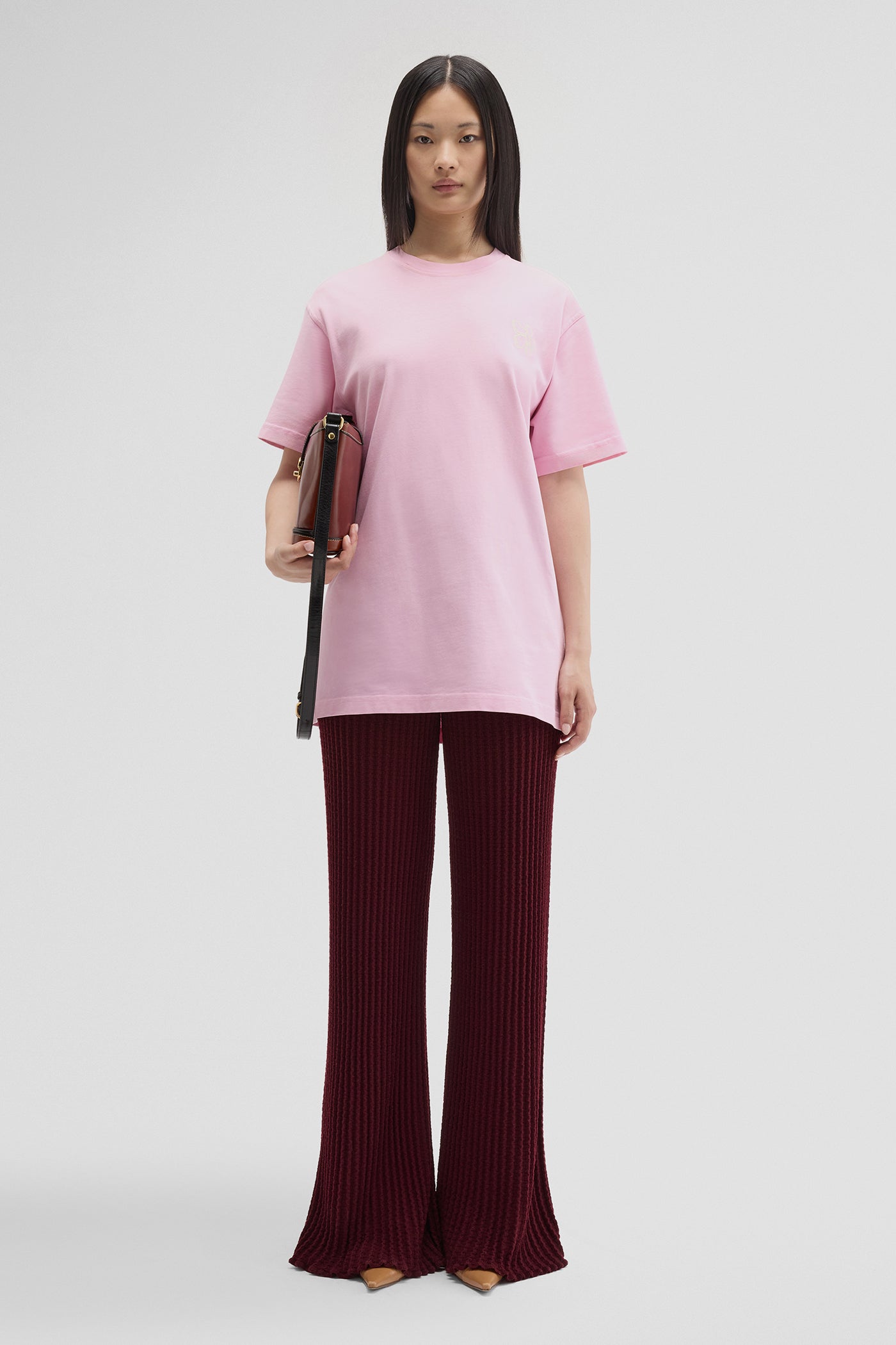 Woman wearing a pink top and burgundy pants holding a brown bag on a white background